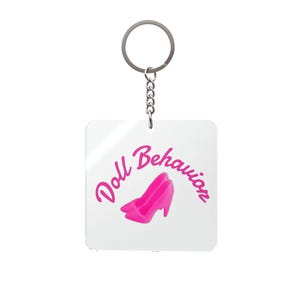 May include: A square keychain with a silver ring and chain. The keychain has the words "Doll Behavior" in pink script, with a pink high-heeled shoe graphic. The background is white.