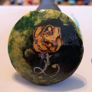 May include: A round, black, ceramic tile with a yellow and orange rose painted on it. The background is a dark green and yellow abstract design.
