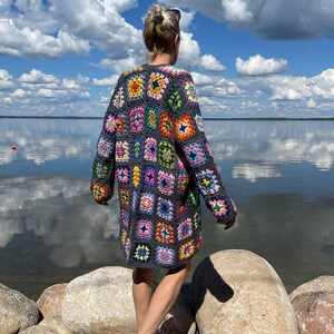 May include: A handmade, long-sleeved crochet cardigan in a patchwork design. The cardigan features a variety of colorful squares in shades of pink, orange, blue, and green, set against a gray background. The person is wearing red sneakers.