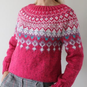 Puede incluir: Un su&eacute;ter de punto rosa con un patr&oacute;n geom&eacute;trico blanco, gris y azul. El su&eacute;ter tiene cuello redondo y mangas largas.