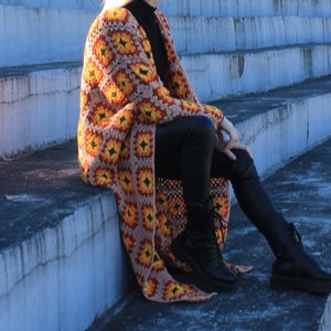 Puede incluir: Una larga chaqueta de punto de ganchillo con un patrón geométrico en tonos marrones, naranjas y amarillos. La chaqueta se lleva sobre pantalones negros y botas negras.