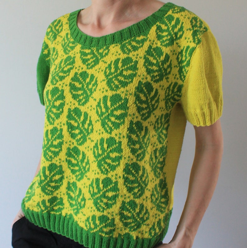 May include: A short-sleeved knitted sweater with a green and yellow monstera leaf pattern. The sweater has a round neckline and a cropped length.