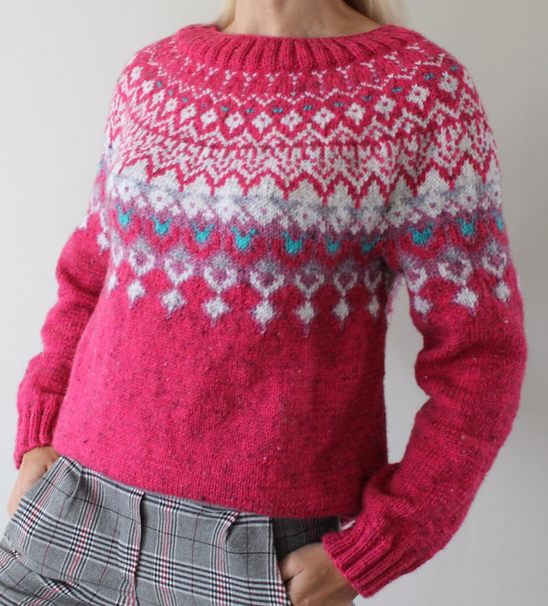 Puede incluir: Un su&eacute;ter de punto rosa con un patr&oacute;n geom&eacute;trico blanco, gris y azul. El su&eacute;ter tiene cuello redondo y mangas largas.