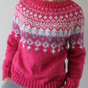 Puede incluir: Un su&eacute;ter de punto rosa con un patr&oacute;n geom&eacute;trico blanco y azul. El su&eacute;ter tiene cuello redondo y mangas largas.