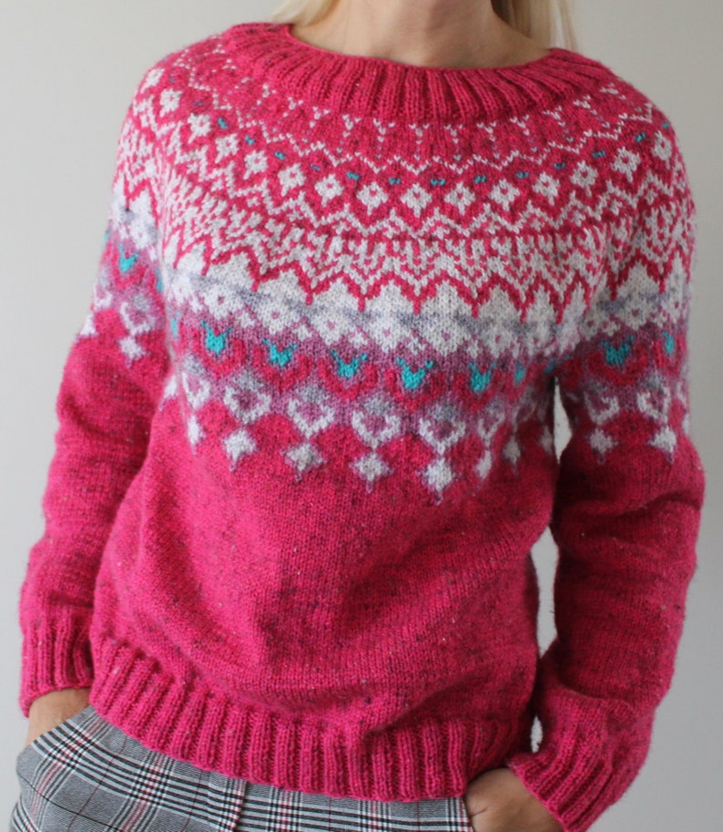 Puede incluir: Un su&eacute;ter de punto rosa con un patr&oacute;n geom&eacute;trico blanco y azul. El su&eacute;ter tiene cuello redondo y mangas largas.