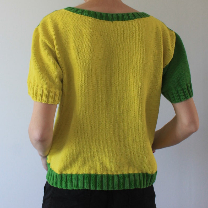 May include: A yellow knitted sweater with green trim around the neckline and sleeves. The sweater is short-sleeved and has a scoop neckline.