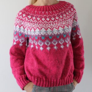 Puede incluir: Un su&eacute;ter de punto rosa con un patr&oacute;n geom&eacute;trico blanco y azul. El su&eacute;ter tiene mangas largas y un cuello redondo.