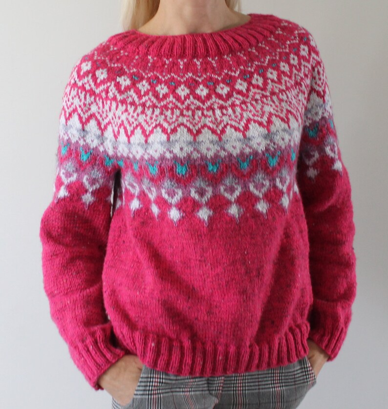 Puede incluir: Un su&eacute;ter de punto rosa con un patr&oacute;n geom&eacute;trico blanco y azul. El su&eacute;ter tiene mangas largas y un cuello redondo.