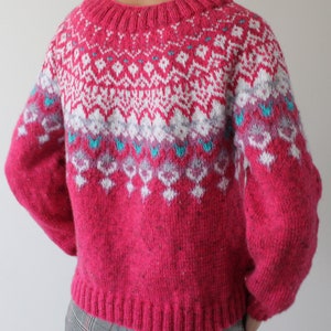 Puede incluir: Un su&eacute;ter de punto rosa con un patr&oacute;n geom&eacute;trico blanco y azul. El su&eacute;ter tiene cuello redondo y mangas largas.