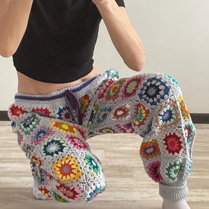 Könnte beinhalten: Handgefertigte Häkelhose mit Patchwork-Design und farbenfrohen Blumenmotiven. Die Hose hat eine graue Basis mit Kordelzug in der Taille und gerippten Bündchen. Die Häkelarbeit enthält verschiedene Farben, darunter Rot, Gelb, Blau und Grün.