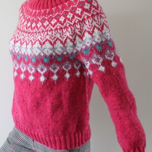 Puede incluir: Un su&eacute;ter de punto rosa con un patr&oacute;n geom&eacute;trico blanco y gris. El su&eacute;ter tiene cuello redondo y mangas largas.