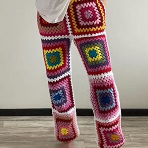 Puede incluir: Pantalones de crochet coloridos con un diseño de patchwork de cuadrados en varios colores, incluyendo rojo, rosa, amarillo, azul y verde. Los pantalones tienen una cintura alta y una pernera acampanada. El trabajo de crochet es detallado e intrincado, mostrando una estética hecha a mano.