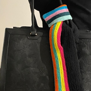 May include: Black tote bag with a floral embossed design. A black knitted sleeve with a rainbow stripe is attached to the bag. The rainbow stripe includes pink, yellow, orange, and blue.