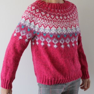 Puede incluir: Un su&eacute;ter de punto rosa con un patr&oacute;n geom&eacute;trico blanco y azul. El su&eacute;ter tiene cuello redondo y mangas largas.