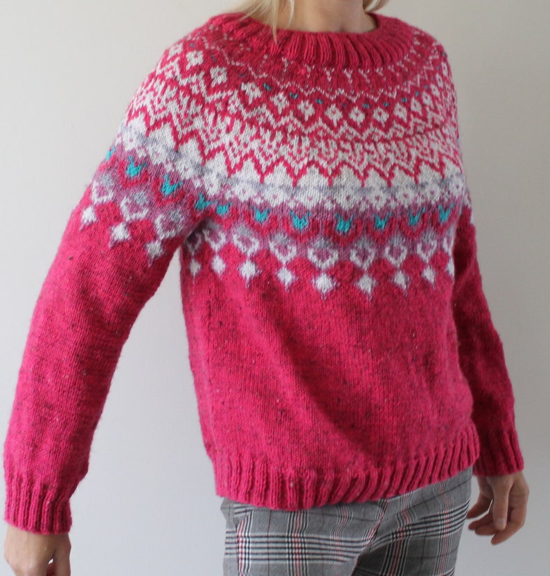 Puede incluir: Un su&eacute;ter de punto rosa con un patr&oacute;n geom&eacute;trico blanco y azul. El su&eacute;ter tiene cuello redondo y mangas largas.