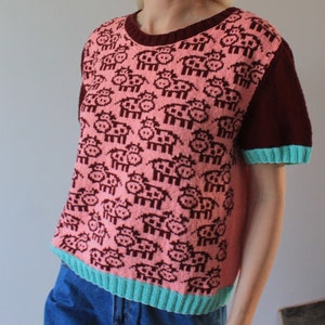 May include: A pink and maroon short-sleeved knitted sweater with a pattern of small pink cows. The sweater has a teal trim around the neckline and sleeves.