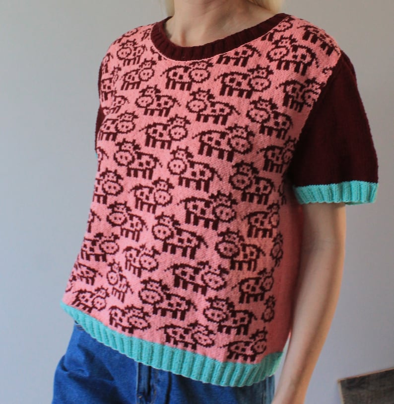 May include: A pink and maroon short-sleeved knitted sweater with a pattern of small pink cows. The sweater has a teal trim around the neckline and sleeves.