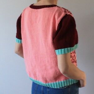 May include: A pink and maroon short-sleeved sweater with a teal trim. The sweater has a knitted design on the sleeves.