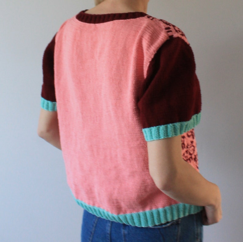 May include: A pink and maroon short-sleeved sweater with a teal trim. The sweater has a knitted design on the sleeves.