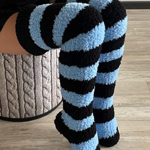 May include: A pair of over-the-knee socks featuring a fuzzy, textured knit pattern with alternating horizontal stripes in black and light blue. The socks have a ribbed cuff at the top and extend from the foot up to the mid-thigh area.