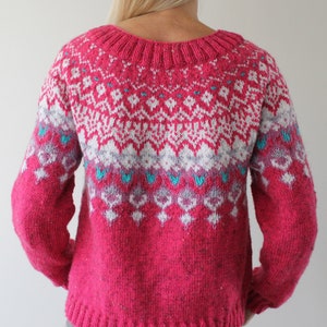 Puede incluir: Un su&eacute;ter de punto rosa con un patr&oacute;n geom&eacute;trico blanco y azul. El su&eacute;ter tiene cuello redondo y mangas largas.