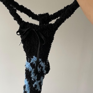 Op de afbeelding: Een zwarte string gemaakt van een pluizig, getextureerd materiaal met een lichtblauw vlamachtig patroon aan de voorzijde. Het kledingstuk heeft een zwart trekkoord in het midden van de taille met zilverkleurige metalen uiteinden.