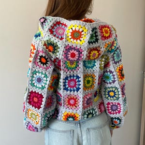 Könnte beinhalten: Graue Häkeljacke mit Kapuze, mit bunten quadratischen Mustern. Jedes Quadrat zeigt ein anderes Blumendesign in Rosa, Gelb, Blau und Grün. Ein einzigartiges, handgefertigtes Kleidungsstück.