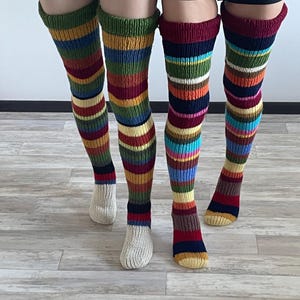 Op de afbeelding: Twee paar kleurrijke, gestreepte dijhoge sokken. Eén paar heeft een groene boord, met gele, rode, blauwe en zwarte strepen. Het andere paar heeft een bordeauxrode boord, met roze, blauwe en bruine strepen. De sokken zijn gemaakt van gebreid materiaal.