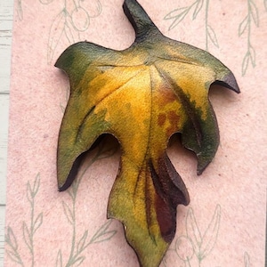 Puede incluir: Un broche de cuero con forma de hoja, en tonos verdes, amarillos y marrones. El broche se muestra en una tarjeta rosa con las palabras "LEATHER PIN" y "WEARABLE NATURE ART".