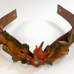 Puede incluir: Una diadema de cuero con un diseño de hojas de otoño en tonos marrones, verdes y naranjas. Las hojas están superpuestas y tienen un aspecto realista.