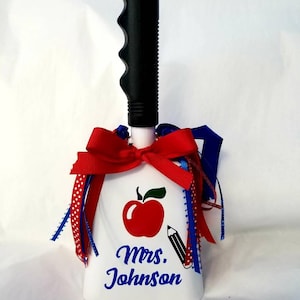 May include: White school bell with a red apple and pencil design. The bell has a black handle and is decorated with red and blue ribbons. The text "Mrs. Johnson" is written in blue on the bell.