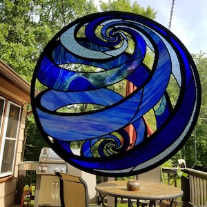 Puede incluir: Una ventana de vitrales colgada de una cadena, con un diseño en espiral azul y blanco. La ventana es circular y tiene un marco negro.