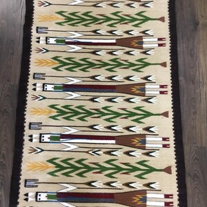 May include: Rectangular beige rug with a repeating pattern of stylized figures and arrows. The figures are in white, brown, and blue, with green arrow designs. The rug has a dark brown border and is made of woven material.