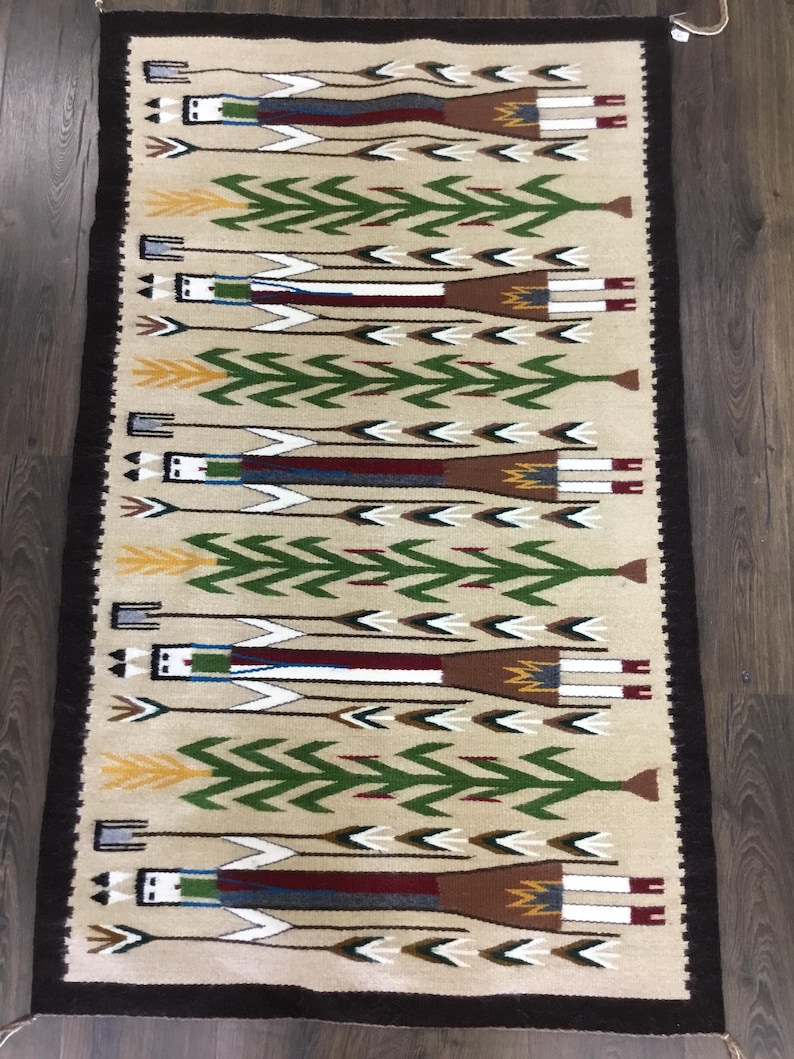 May include: Rectangular beige rug with a repeating pattern of stylized figures and arrows. The figures are in white, brown, and blue, with green arrow designs. The rug has a dark brown border and is made of woven material.