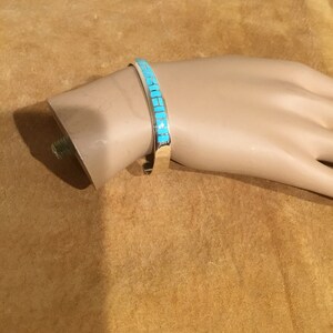 May include: A silver bracelet with a turquoise inlay design. The bracelet is on a brown surface.