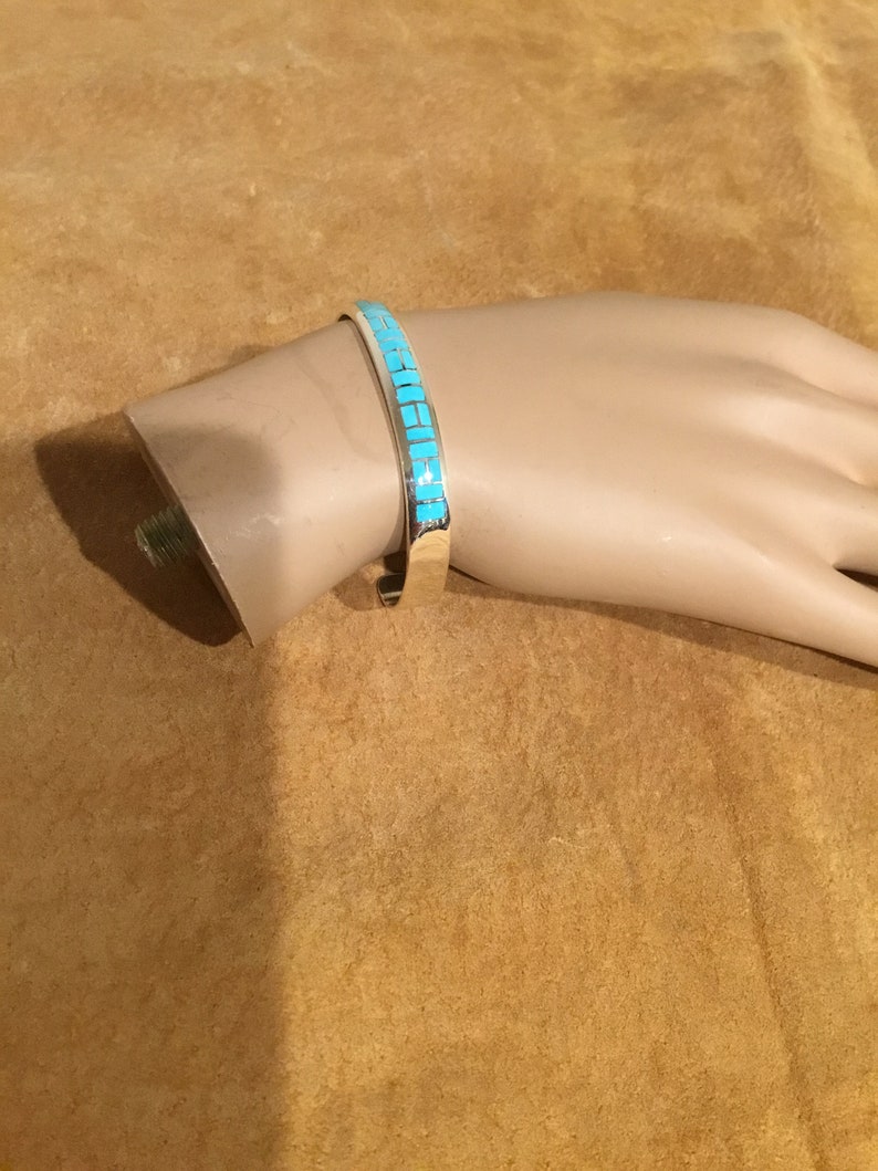 May include: A silver bracelet with a turquoise inlay design. The bracelet is on a brown surface.