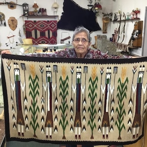 May include: A beige woven rug with a dark brown border. The rug features a repeating pattern of stylized figures with green, brown, white, and blue elements, interspersed with green stalks. The rug is held up by a person.