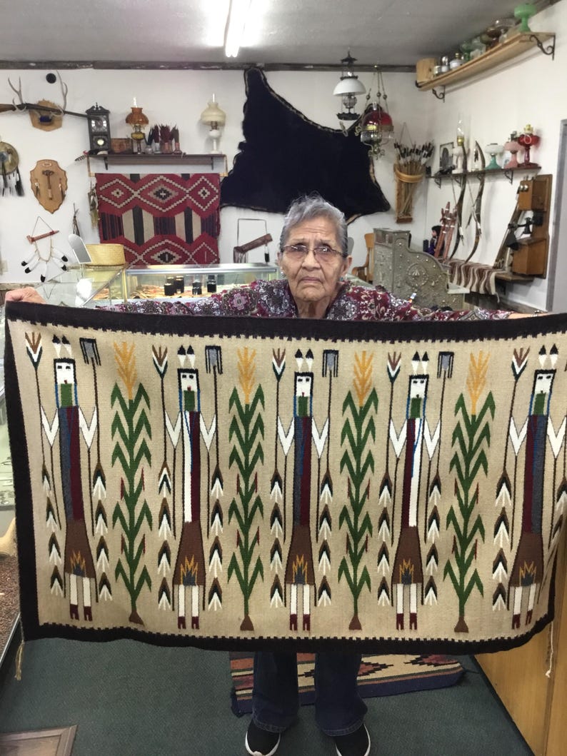 May include: A beige woven rug with a dark brown border. The rug features a repeating pattern of stylized figures with green, brown, white, and blue elements, interspersed with green stalks. The rug is held up by a person.