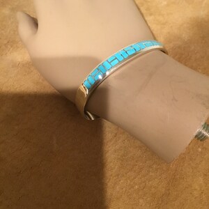 May include: A silver bracelet with a turquoise inlay design. The bracelet is on a mannequin's wrist.