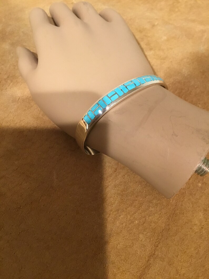 May include: A silver bracelet with a turquoise inlay design. The bracelet is on a mannequin's wrist.