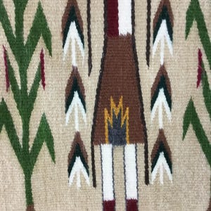May include: Close-up of a woven textile with a beige background. The design features a central figure in brown with white legs and red feet, surrounded by stylized trees and arrow-like shapes in green, white, and brown. The border is a dark brown.