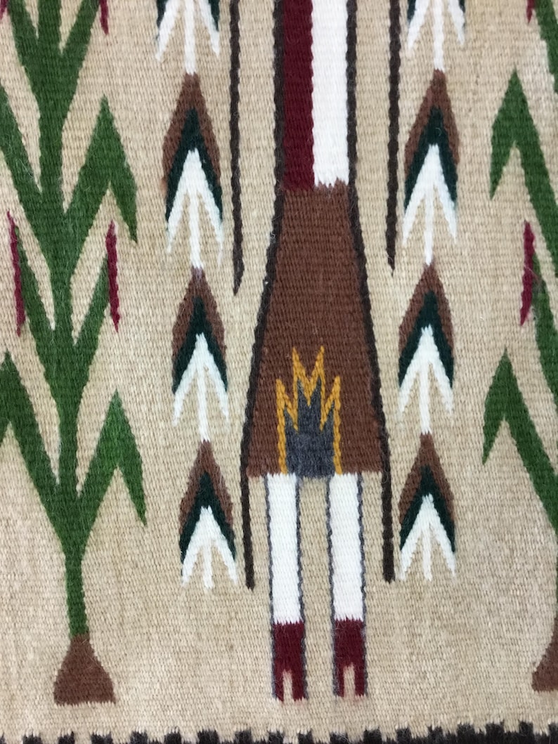 May include: Close-up of a woven textile with a beige background. The design features a central figure in brown with white legs and red feet, surrounded by stylized trees and arrow-like shapes in green, white, and brown. The border is a dark brown.
