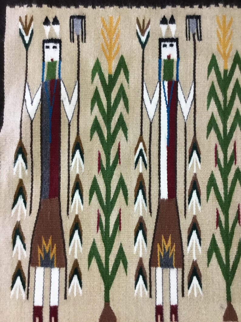 May include: A woven textile featuring two stylized figures with blue, green, red, and brown details. The figures are flanked by green and yellow plant motifs, all set against a beige background. The design has a Native American aesthetic.