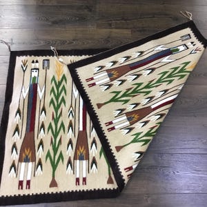 May include: Two rectangular woven rugs with a beige background and a dark brown border. One rug is partially folded over, revealing a detailed pattern of stylized figures, arrows, and geometric shapes in various colors, including green, red, and brown.