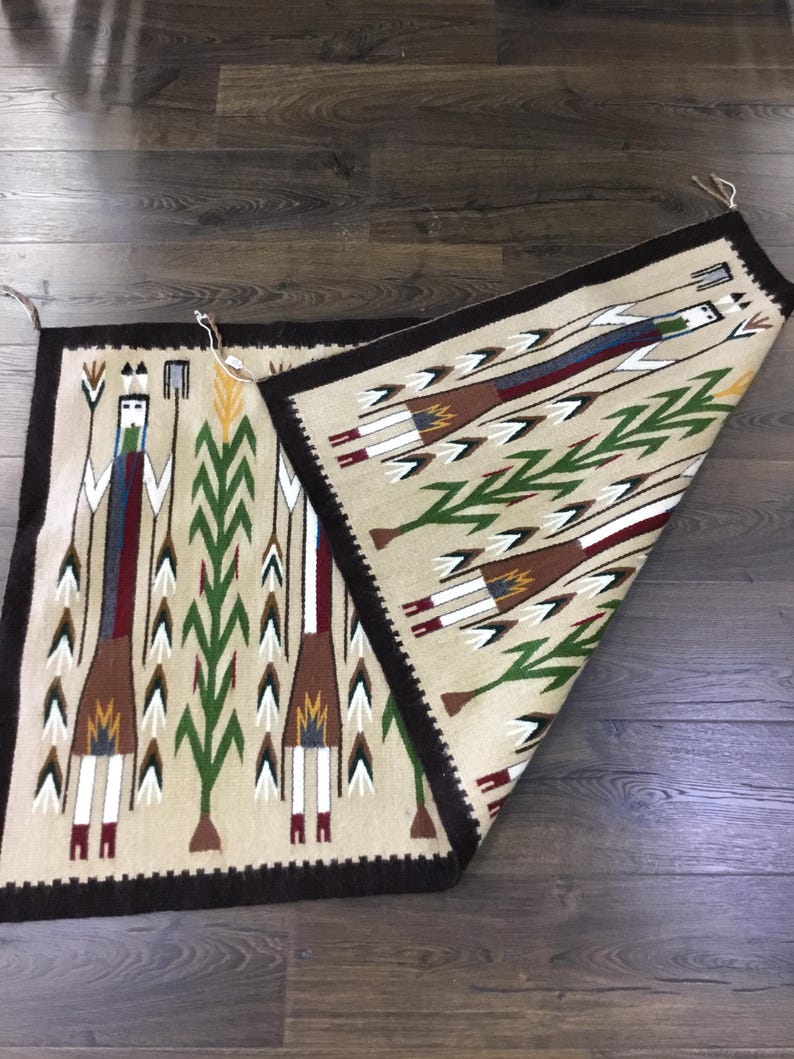 May include: Two rectangular woven rugs with a beige background and a dark brown border. One rug is partially folded over, revealing a detailed pattern of stylized figures, arrows, and geometric shapes in various colors, including green, red, and brown.