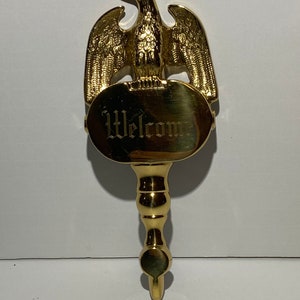 May include: Gold-toned metal welcome sign with an eagle perched on top. The sign is oval shaped and has the word "Welcome" engraved on it.