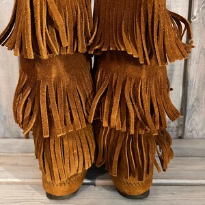 May include: A pair of brown suede boots with multiple layers of fringe. The boots have a moccasin-style design and a low heel. The fringe adds a decorative element to the boots.
