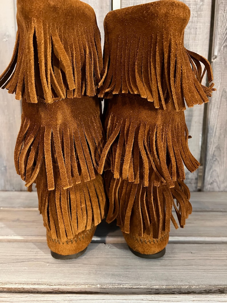 May include: A pair of brown suede boots with multiple layers of fringe. The boots have a moccasin-style design and a low heel. The fringe adds a decorative element to the boots.