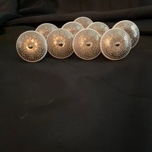 May include: A collection of ten decorative cabinet knobs. Each knob features a silver-colored, patterned face with a central, small, silver-colored stud. The knobs have a white base and are arranged on a black surface.