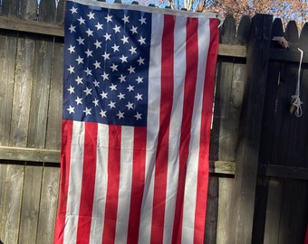 May include: An American flag hanging on a wooden fence. The flag is red, white, and blue with 13 stripes and 50 stars.